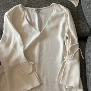 Vit blus med knytband - Elegant vit blus med v-ringning och långa ärmar. Blusen har dekorativa knytband vid ärmsluten som ger en stilfull touch. Perfekt för en chic och avslappnad look.