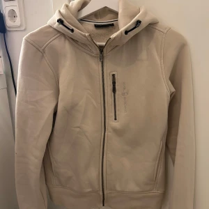 Beige huvtröja från Sail Racing - Säljer en stilren beige huvtröja från Sail Racing med dragkedja framtill och en praktisk ficka med dragkedja på bröstet. Tröjan har en bekväm huva med justerbara snören och långa ärmar. Perfekt för en avslappnad look.