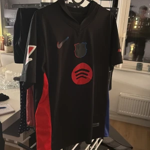 Barcelona away kit , Lamine yamal  - Säljer en svart fotbollströja från Nike med korta ärmar. Tröjan har ett klubbmärke på bröstet och röda detaljer, inklusive texten 'Lamine Yamal' och nummer 19 på ryggen. Tillverkad i Dri-FIT-material för optimal komfort.
