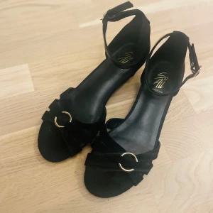 Svarta sandaler med gulddetaljer - Eleganta svarta sandaler med guldfärgade spännen och remmar runt ankeln. Perfekta för sommarens alla tillfällen.