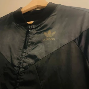 Svart bomberjacka från Adidas - Snygg svart bomberjacka från Adidas med dragkedja och ribbad krage. Jackan har en stilren design med det klassiska Adidas-loggan på bröstet. Perfekt för en sportig look.