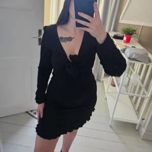 Svart strukturerad klänning med volangdetaljer - Snygg svart klänning med volangdetaljer och knyt framtill. Den har en v-ringad design och långa ärmar. Perfekt för en stilren look. Använd ett nyår.