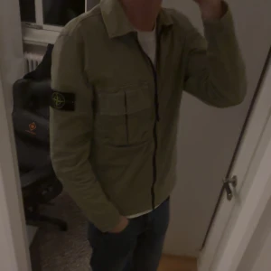 Olivgrön overshirt från Stone Island - Säljer en stilren olivgrön overshirt från Stone Island. Jackan har en dragkedja framtill och en klassisk krage. Den har en ficka på bröstet och den ikoniska Stone Island-loggan på ärmen. Perfekt för lager-på-lager-stil. Enstaka fläck på höger armen men inte något som inte går att tvätta bort👍