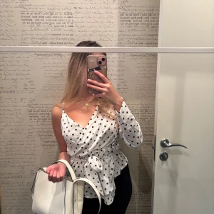 Vit prickig omlottblus - Säljer en stilren vit omlottblus med svarta prickar. Blusen har en knytning i midjan. NA-KD 140kr