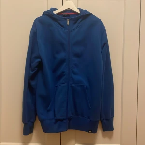 Blå hoodie med dragkedja - Säljer en snygg blå hoodie med dragkedja och huva. Tröjan har långa ärmar och två praktiska fickor framtill. Perfekt för en avslappnad stil.