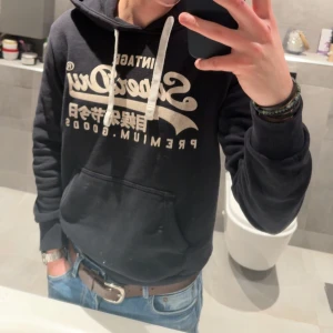 Svart hoodie från Superdry - Säljer en svart hoodie från Superdry med tryck framtill. Den har en justerbar huva med vita snören och en stor magficka. Perfekt för en avslappnad stil.