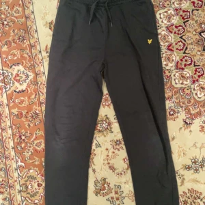 Svarta mjukisbyxor från Lyle & Scott - Säljer ett par svarta mjukisbyxor från Lyle & Scott med elastisk midja och snörning. Byxorna har en gul logga på framsidan och en bakficka. Perfekta för en avslappnad stil.
