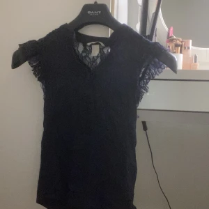 Mörkblå spetsdetaljerad topp från h&m - Snygg mörkblå topp från h&m med spetsdetaljer vid ärmarna och halsen. Toppen har en v-ringning och är ärmlös men går att knäppa uppe, vilket ger en elegant look. Perfekt för att klä upp en outfit!