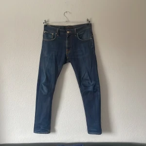 Blå jeans från Nudie Jeans - Snygga blå jeans från Nudie Jeans med klassisk femficksdesign och knappgylf. De har en slim passform och är perfekta för en stilren look. Märkesdetaljer syns på baksidan.