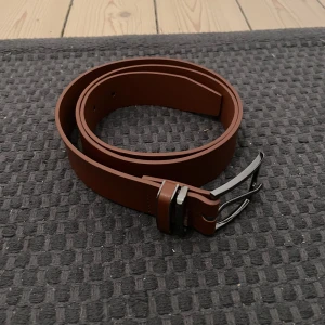 Brunt läder bälte - Snyggt brunt läder skärp från Pier One med en stilren silverfärgad spänne. Perfekt för att ge en klassisk touch till din outfit. Skärpet har flera hål för justerbar passform.