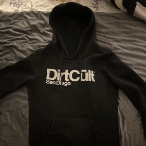 Svart hoodie från DirtCült - Säljer en svart hoodie från DirtCült med texten 'San Diego' tryckt på framsidan. Den har en klassisk känguruficka och justerbar huva med dragsko. Perfekt för en avslappnad stil.