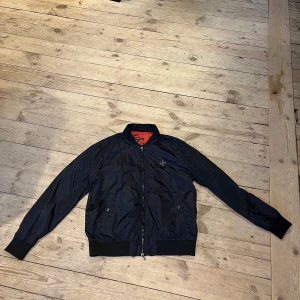 Svart bomberjacka från J.Lindeberg - Snygg svart bomberjacka från J.Lindeberg med dragkedja och ribbade muddar. Jackan har en klassisk design med en liten logotypdetalj på bröstet och en kontrasterande orange insida. Perfekt för en stilren look. Använd fåtalet gånger. Pris går att diskutera vid snabb affär 🤝