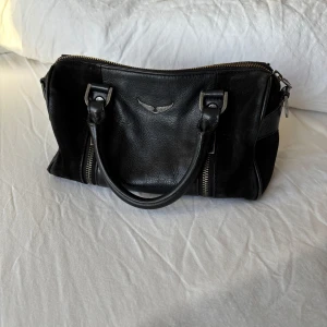 Zadig väska  - Snygg svart Zadig handväska i skinn med dragkedjor och metallspännen. Perfekt för att ge en stilren touch till din outfit. Väskan har två handtag och en praktisk storlek. Använt skick, defekt på dragkedja som tappat färg samt på två ställen där tyget har lossnat. Går säkert att laga.                Längd: 23 cm. Höjd: 14 cm.