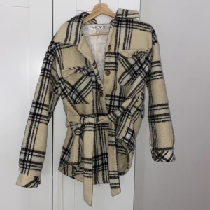 Beige rutig jacka från Missguided - Säljer en snygg beige rutig jacka från Missguided. Jackan har långa ärmar och ett bälte i midjan för en schysst passform. Den är försedd med knappar framtill och har en mysig känsla tack vare sitt mjuka material.