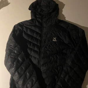Svart dunjacka från Haglöfs - Säljer en svart dunjacka från Haglöfs med huva och dragkedja. Jackan är quiltad och har en stilren design. Dragkedjorna på fickorna är sönder men allt annat är som det ska. 