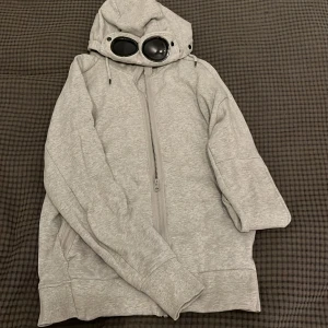 Cp company hoodie - Säljer en unik grå huvtröja från C.P. Company med inbyggda glasögon i huvan. Tröjan har dragkedja framtill och långa ärmar. Perfekt för en cool och avslappnad stil. 