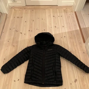 Svart dunjacka från Peak Performance - Säljer en svart dunjacka från Peak Performance. Jackan har en dragkedja framtill och en huva för extra skydd mot väder och vind. Perfekt för kyligare dagar.