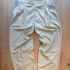 Beige kostymbyxor från Doni - Snygga beige kostymbyxor från Doni med klassisk design. Byxorna har en relativt baggy passform och är perfekta för en stilren look. De har knappar och dragkedja framtill samt en bakficka med knapp. Perfekta för en elegant och sofistikerad stil. Storleken är ungefär 34/32. Skriv vid frågor!