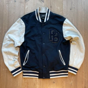 Svart och vit bomberjacka från Pull&Bear - Säljer en snygg mörkblå och vit bomberjacka från Pull&Bear. Jackan har vita ärmar och mörkblåa detaljer med broderad logga på bröstet. Den har knappar framtill och randiga muddar vid ärmslut och krage. Perfekt för en cool och avslappnad stil. Använd fåtal gånger, skriv vid frågor!