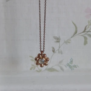 Halsband med blomformad berlock - Elegant halsband med en blomformad berlock i guldton. Berlocken har en glittrande sten i mitten som ger en fin detalj. Kedjan är tunn och justerbar, vilket gör det enkelt att anpassa längden. Perfekt för att lägga till en subtil touch av glamour till din outfit. Baksidan på "blomman" syns att den varit använd men framsidan är bra skick. 