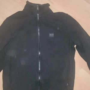 Svart jacka från BOSS - Säljer en stilren svart jacka från BOSS med dragkedja framtill. Jackan har en hög krage och ett diskret BOSS-märke på bröstet. Perfekt för dig som vill ha något simpelt! Den är använd 1 gång! 