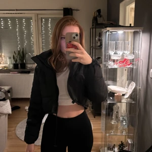 Svart croppad pufferjacka - Säljer en svart croppad pufferjacka med dragkedja och snörning i nederkant. Jackan har långa ärmar och en hög krage, perfekt för kyligare dagar. Den är både stilren och trendig, idealisk för att hålla värmen med stil.