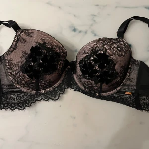 Svart spets-bh med rosett och pärldetalj - Elegant svart bh i spets med vacker rosett och pärldetalj mellan kuporna. Den har justerbara axelband och en fin spetskant runtom. Perfekt för en lyxig känsla.