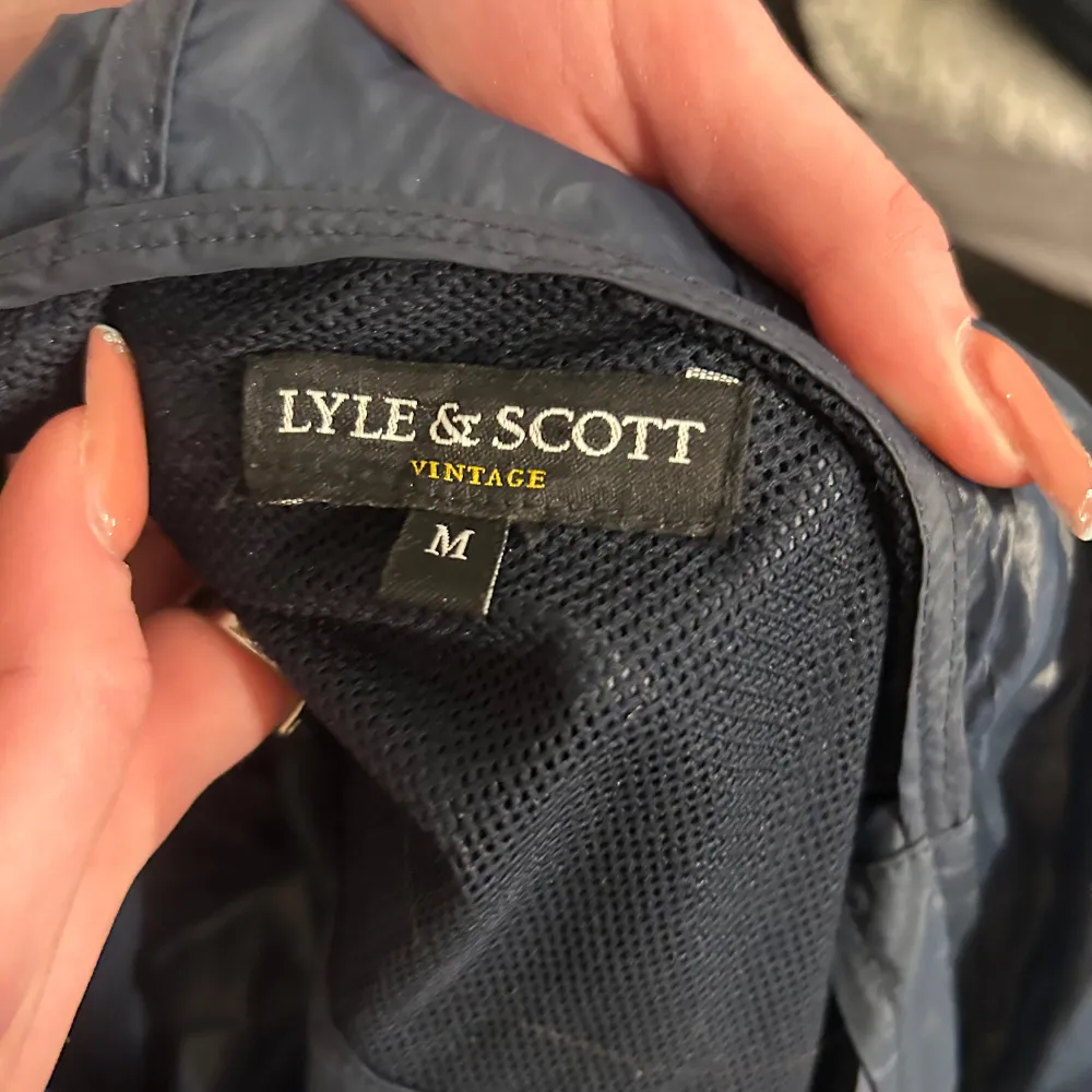 Säljer en snygg blå vindjacka från Lyle & Scott med dragkedja och huva. Jackan har ett gult broderat emblem på bröstet och justerbara snören vid huvan. Perfekt för blåsiga dagar!. Takit.