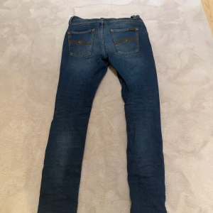 Blå jeans från Nudie Jeans - Snygga blå jeans från Nudie Jeans med klassisk femficksdesign och orange sömmar. Jeansen har en normal passform och är tillverkade i slitstarkt denim. Perfekta för en avslappnad stil.