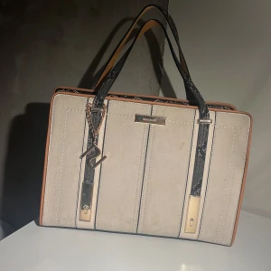 Beige och brun handväska från River Island - Snygg handväska från River Island i beige och brun med svarta detaljer. Väskan har ett elegant ormskinnsmönster på handtagen och insidan. Perfekt för att bära dina dagliga nödvändigheter med stil. Bredd: 34cm  Höjd: 24cm Djup: 14,5 cm 