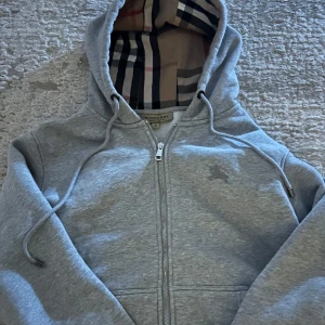 Grå hoodie från Burberry - Säljer en stilren grå hoodie från Burberry med dragkedja och klassiskt rutigt mönster i huvan. Perfekt för en avslappnad look. Den har långa ärmar och justerbara snören vid huvan.