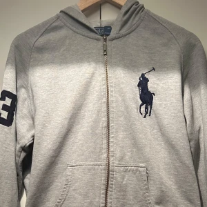 Grå hoodie från Ralph Lauren - Säljer en stilren grå hoodie från Ralph Lauren med dragkedja och stor logga på bröstet. Tröjan har en broderad siffra på ärmen och praktiska fickor framtill. Perfekt för en avslappnad look.