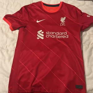 Liverpool fotbollströja från Nike - Säljer en röd Liverpool fotbollströja från Nike med korta ärmar och snygga diagonala ränder. Tröjan har klubbens emblem och sponsorlogga på framsidan. Perfekt för alla Liverpool-fans! 🔴 nypris: 1100