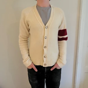Vit/Beige kofta med röda ränder - Säljer en snygg beige/vit kofta med knappar och röda ränder på ärmen. Perfekt för kyliga dagar och ger en stilren look. 