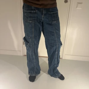 Blå cargobyxor från BDG - Säljer ett par blå cargobyxor från BDG med flera praktiska fickor och en avslappnad passform. Byxorna har en cool Y2K-stil och är perfekta för en casual look. De är tillverkade i jeansmaterial och har en knapp och dragkedja framtill.