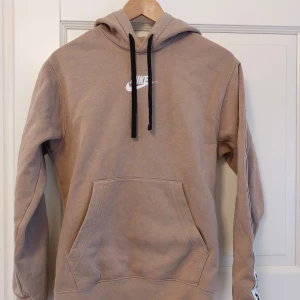 Beige hoodie från Nike - Säljer en snygg beige hoodie från Nike med svart dragsko och vit logga på bröstet samt detaljen Nike-loggor längs ärmarna. Perfekt för en avslappnad stil.
