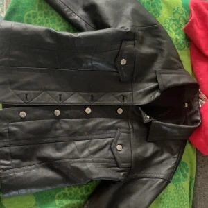 Svart skinnjacka med knappar - Snygg svart skinnjacka med knappar och stilrena detaljer. Jackan har en klassisk krage och långa ärmar. Perfekt för en cool look.