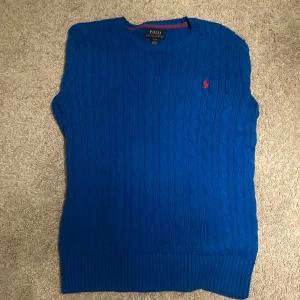 Blå långärmad/kabelstickad tröja från Polo Ralph Lauren - Snygg blå långärmad tröja från Polo Ralph Lauren med kabelstickat mönster och röd logga på bröstet. Perfekt för lager-på-lager-stil eller sommarens varma dagar. (Pris kan diskuteras)