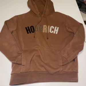 Brun hoodie från Hoodrich - Säljer en snygg brun hoodie från Hoodrich med texten 'HOODRICH' i svart, beige och vitt på bröstet. Hoodien har en klassisk känguruficka och justerbar huva med snören. Perfekt för en avslappnad stil.