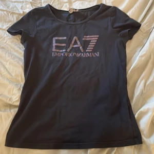 Svart t-shirt från Emporio Armani - Snygg svart t-shirt från Emporio Armani med EA7-logga på framsidan. Den har en rund halsringning och korta ärmar, perfekt för en stilren look. Passar bra till både jeans och kjol. 