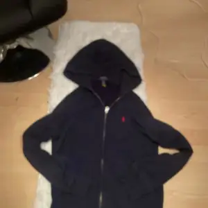 Säljer en snygg mörkblå hoodie från Polo Ralph Lauren. Den har en dragkedja framtill och en klassisk röd logga på bröstet. Perfekt för kyliga dagar och ger en stilren look. Passar både till vardags och lite mer avslappnade tillfällen. Barn storlek L