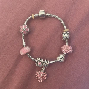 Charmigt armband med rosa detaljer - Säljer ett superfint armband med flera olika berlocker. Armbandet är i silverfärg och har söta rosa detaljer. Det finns en blomma, ett hjärta och en glittrig hjärtberlock som verkligen sticker ut. Perfekt för att ge lite extra bling till din outfit!