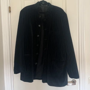 Svart randig kavaj - Säljer en stilren svart sammet kavaj med tunna vita ränder. Kavajen har en klassisk design med knappar framtill och fickor. Perfekt för både formella och semi-formella tillfällen. Passar bra till både vår och höst.