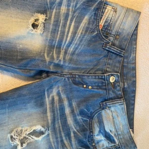 Blå slitna jeans från Diesel - Säljer ett par coola blå jeans från Diesel med slitningar och hål för en trendig look. De har en normal passform och är perfekta för en avslappnad stil. Jeansen har klassiska fem fickor och en knappgylf. Perfekta för både vardag och fest!
