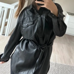 Svart skinnklänning från Zara - Snygg svart skinnjacka från Zara med långa ärmar och knappar framtill. Den har en cool bälte i midjan för en perfekt passform. Perfekt för höst och vinter, och funkar lika bra till fest som till vardags. 🖤knappt använd 