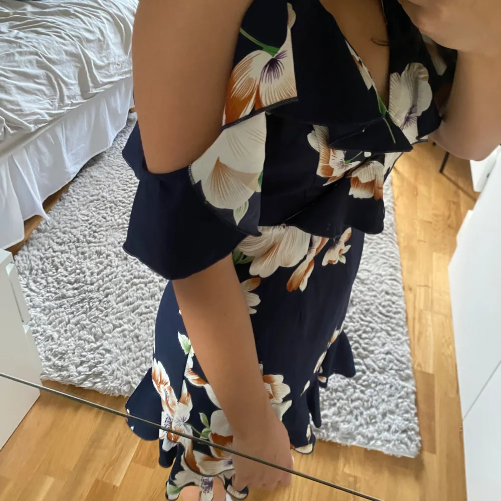 Säljer en superfin mörkblå klänning med stora blommor i vitt och orange. Den har en omlott-design och offshoulder-ärmar som ger en romantisk look. Perfekt för sommarens fester eller en mysig vårdag. Klänningen är kort och har en volangdetalj vid fållen.. Mekot.