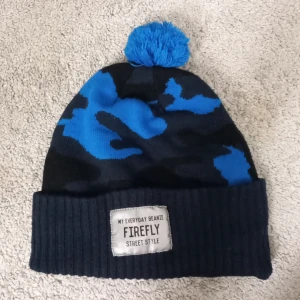 Blå och svart mössa från Firefly - Säljer en cool mössa från Firefly i blått och svart med ett camouflagemönster. Den har en ribbad kant och en blå tofs på toppen. Perfekt för att hålla sig varm med stil under kalla dagar. Märket 'My Everyday Beanie' ger den en extra touch av street style. Är däremot nästan helt ny och pris 120kr