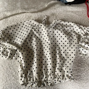 Vit prickig topp från Boohoo - Säljer en superfin vit topp med svarta prickar från Boohoo. Den har korta ärmar med volangdetaljer och en söt knytning i nacken. Toppen är croppad och har en elastisk kant nedtill för en bekväm passform. Perfekt för en somrig look! Använd några få gånger.