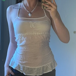 Vit topp med fjäril och pärlor - Denhär toppen är köpt vintage. Den har en fjäril på bröstet samt pärldetaljer. I ryggen går axelbandet i kors. Den har ett litet hål vid höger bröst, men det synns inte. Annars är den i fint skick utan skador. Skriv privat om ni har frågor 🤍