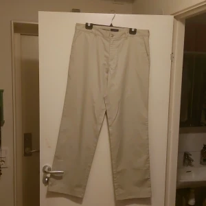 Beige byxor från Atlant - Säljer ett par stilrena beige byxor från Atlant. De har en klassisk design med raka ben och är tillverkade i ett bekvämt material. Byxorna har knappar bak och en midwaist passform, perfekt för både vardag och lite mer uppklädda tillfällen.